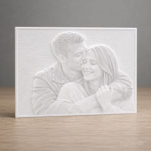 Può includere: Una cornice fotografica bianca stampata in 3D con una coppia che si abbraccia e si bacia. L'immagine è una scultura in rilievo, con figure e sfondo strutturati. La cornice è rettangolare e poggia su una superficie di legno.