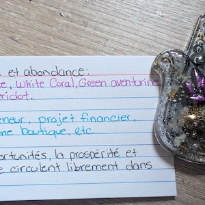 May include: A clear resin Hamsa hand with glitter and a purple flower design. A handwritten note in French lists crystals and affirmations for abundance and opportunity.