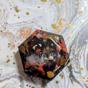 Op de afbeelding: Een donker, zeshoekig kunstwerk van hars met ingebedde stenen, goudvlokken en andere materialen. Het stuk heeft een mix van rode, oranje, bruine en gele elementen, met een heldere, glanzende afwerking. De achtergrond is een gemarmerd patroon met gouden accenten.