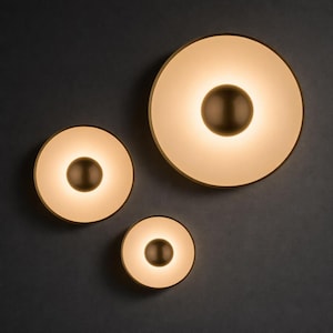 Könnte beinhalten: Drei moderne Wandleuchten mit kreisförmigem Design. Jede Leuchte hat eine warme, leuchtende Mitte, einen weißen Ring und einen bronzefarbenen Außenrand. Sie sind an einer dunkelgrauen Wand montiert.
