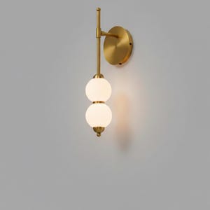 May include: A modern wall sconce with a brass finish. The fixture features two spherical, frosted glass shades stacked vertically. The sconce is mounted on a circular brass base and has a slender, vertical arm.
