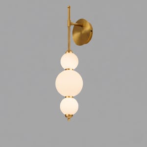 May include: A modern wall sconce featuring three frosted glass globes of varying sizes, stacked vertically. The fixture has a brass finish, including the mounting plate and connecting rod. The design is minimalist and elegant, suitable for interior lighting.