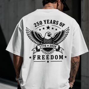 May include: A white short-sleeve t-shirt featuring a black graphic on the back. The design displays a bald eagle with spread wings above a banner reading 1776-2026, surrounded by the text 250 YEARS OF FREEDOM and several small star icons.