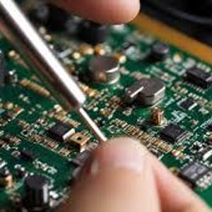 以下が含まれることがあります： さまざまな電子部品を備えた緑色の回路基板のクローズアップ。人がはんだごてを使って基板をいじっています。基板には金と銀色のコンポーネントがあります。