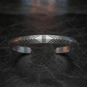 Puede incluir: Una pulsera de plata con diseño abierto. La pulsera presenta un patrón geométrico de flechas y líneas. El metal tiene un acabado cepillado. La pulsera se muestra sobre una superficie oscura y texturizada.