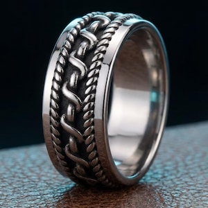 Puede incluir: Un anillo plateado con un diseño trenzado detallado. El anillo presenta una banda central de patrones entrelazados, bordeada por una textura similar a una cuerda. El anillo se muestra sobre una superficie marrón con un fondo oscuro.