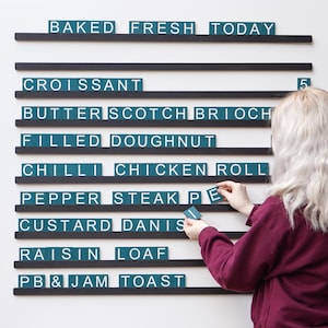 May include: A bakery menu board with black shelves and teal letter tiles. The menu includes items like croissant, filled doughnut, and pepper steak pie. A person is arranging the letters on the board.