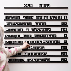May include: A black and white menu board with the words "OUR MENU" at the top. The menu lists breakfast and lunch items like toast, waffles, and sandwiches. A person is adjusting the letters on the board.