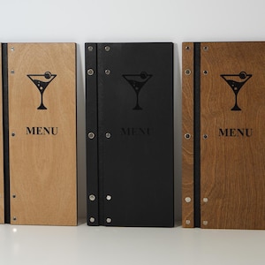 May include: Three rectangular wooden menu covers with a vertical black spine secured by silver metal rivets. Each cover features a black laser-engraved martini glass icon above the word MENU. The set includes one light wood, one black, and one dark wood.