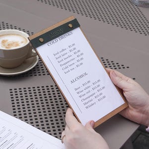 May include: A rectangular wooden menu board with a dark green leather-like top clip secured by two gold-toned metal rivets. The white paper insert features the text MENU, COLD DRINKS, and ALCOHOL with a list of beverage items and their prices.