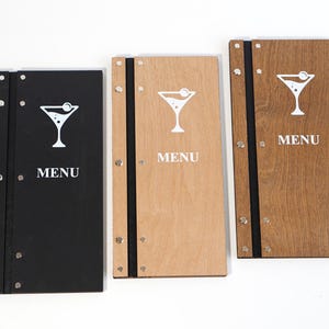 Könnte beinhalten: Drei hölzerne Menüumschläge in Schwarz, Beige und Braun. Jeder Umschlag zeigt ein weißes Cocktailglas-Logo und das Wort "MENU" in Weiß. Die Umschläge sind mit Metallnieten und einem schwarzen Gummiband gebunden.