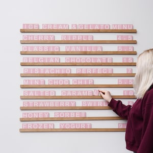 May include: A menu board made of wood with pink letter tiles, listing ice cream and gelato flavours. Flavours include Cherry Ripple, Classic Vanilla, and Belgian Chocolate. A person is seen adjusting the menu.
