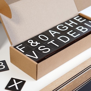 Puede incluir: Un juego de Scrabble con fichas negras y letras blancas, presentado en una caja de cartón abierta. La caja contiene las fichas, con fichas sueltas adicionales y atriles de madera visibles. La caja tiene el texto "W4X4Y4Z4" y "18 2 5 3 5 4 5 5 8 6 5 7 5 8 59".