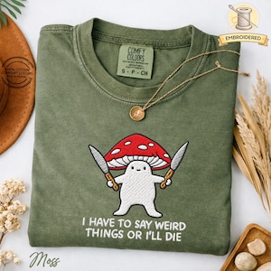 May include: Moss green t-shirt with an embroidered mushroom character holding knives, and the text "I HAVE TO SAY WEIRD THINGS OR I'LL DIE." A gold necklace with a pendant rests on the shirt. The shirt is labeled "COMFY COLORS."