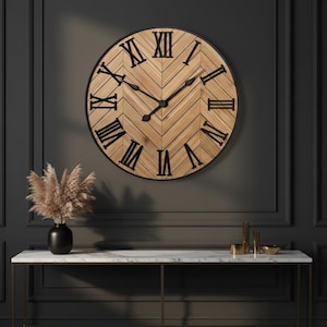 May include: A large, round wall clock with a light brown, herringbone-patterned face and black Roman numerals. The clock is framed in black and hangs on a dark gray wall. The clock's hands are black.