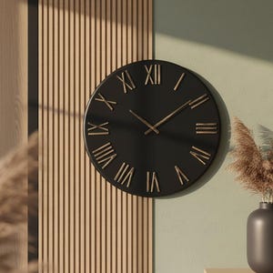 May include: A round black wall clock with gold Roman numerals and hands. The clock is mounted on a light green wall next to a wooden panel. A vase with dried plants sits below the clock.