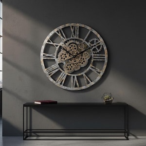 May include: A large, round wall clock with a gear mechanism design. The clock features Roman numerals and a distressed metal finish. The clock is mounted on a gray wall above a black console table. A geometric terrarium and a book are on the table.