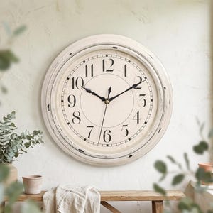 May include: A round wall clock with a distressed off-white frame and a cream-colored face. The clock features black Arabic numerals from one to twelve, a thin black minute track, and two black metal hands centered on the dial.