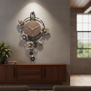 May include: A decorative wall clock with a brown face and gold hands. The clock is surrounded by a black metal frame with floral accents. The clock is mounted on a gray wall above a wooden cabinet.