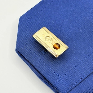 May include: A rectangular gold-tone metal cufflink featuring a smooth surface with an engraved swirl design and a small, round, brown tiger's eye gemstone inlay. The cufflink is attached to the corner of a vibrant blue fabric shirt cuff.