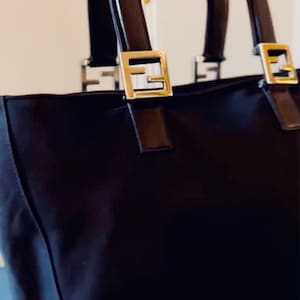 Op de afbeelding: Zwarte tote bag met bruine handvatten en goudkleurige metalen accenten. De tas heeft een rechthoekige vorm met een gestructureerd ontwerp. De handvatten zijn gebogen en bevestigd met goudkleurige metalen hardware.