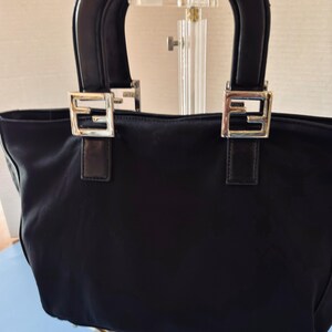 Op de afbeelding: Zwarte tote bag met twee korte handvatten. De tas heeft zilverkleurige vierkante gespen met een gestileerd "F"-ontwerp aan de voorkant. De handvatten zijn zwart en de tas lijkt gemaakt van een nylonachtig materiaal.