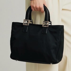 Op de afbeelding: Zwarte tote bag met zilverkleurige hardware. De tas heeft een rechthoekige vorm met twee handvatten aan de bovenkant en een stoffen body. De handvatten zijn bevestigd met zilverkleurige metalen hardware. De tas wordt door een persoon vastgehouden.