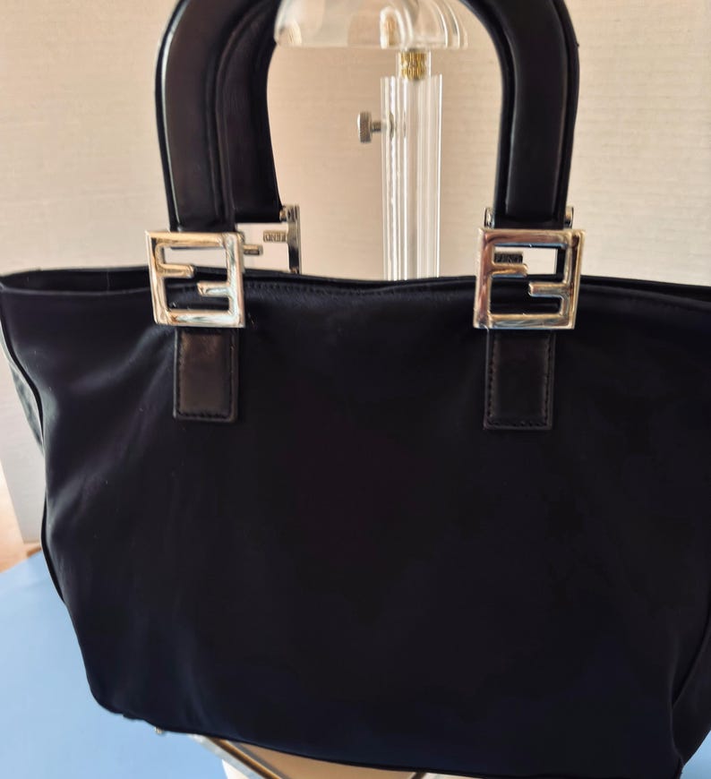 Op de afbeelding: Zwarte tote bag met twee korte handvatten. De tas heeft zilverkleurige vierkante gespen met een gestileerd "F"-ontwerp aan de voorkant. De handvatten zijn zwart en de tas lijkt gemaakt van een nylonachtig materiaal.