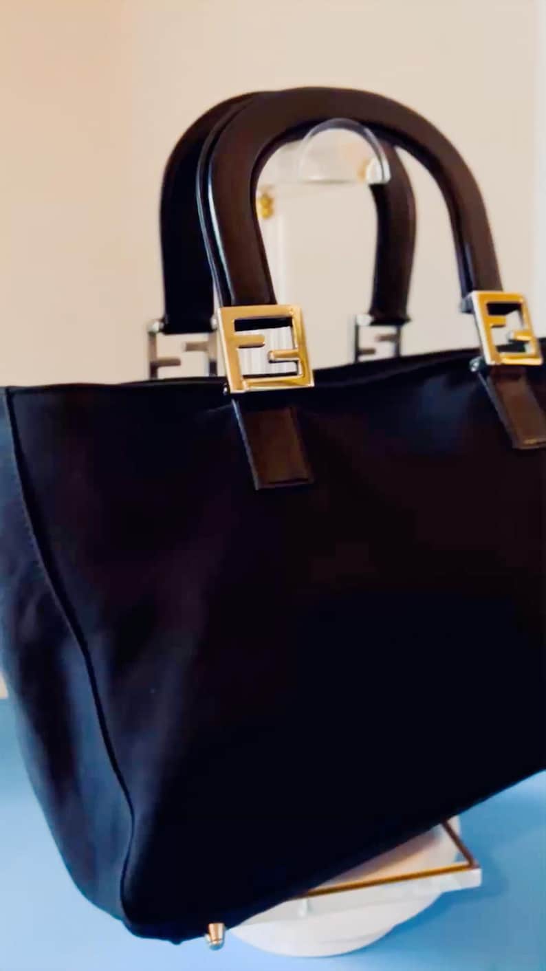 Op de afbeelding: Zwarte tote bag met bruine handvatten en goudkleurige metalen accenten. De tas heeft een rechthoekige vorm met een gestructureerd ontwerp. De handvatten zijn gebogen en bevestigd met goudkleurige metalen hardware.