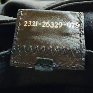 Op de afbeelding: Close-up van een donkerbruin leren label met gouden tekst. De tekst luidt "2321-26329-079". Het label is gestikt op een zwarte stof. Het label is rechthoekig met afgeronde hoeken.