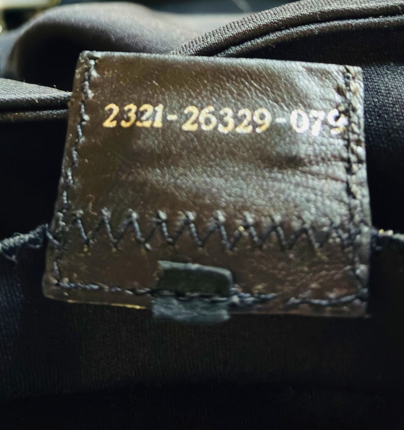 Op de afbeelding: Close-up van een donkerbruin leren label met gouden tekst. De tekst luidt "2321-26329-079". Het label is gestikt op een zwarte stof. Het label is rechthoekig met afgeronde hoeken.