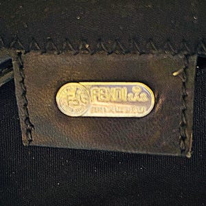 Op de afbeelding: Close-up van een zwart leren label met een zilveren Fendi-logo. Het ovale logo toont de merknaam en de tekst 'Made in Italy'. Het label is gestikt op een zwarte stof.