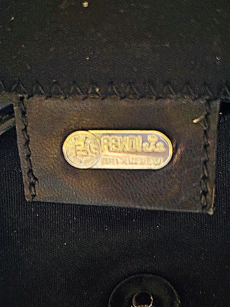 Op de afbeelding: Close-up van een zwart leren label met een zilveren Fendi-logo. Het ovale logo toont de merknaam en de tekst 'Made in Italy'. Het label is gestikt op een zwarte stof.