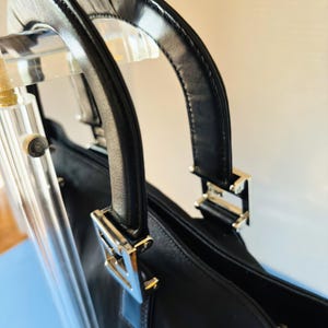 Op de afbeelding: Zwarte handtas met twee afgeronde leren handvatten en zilverkleurige metalen hardware. De handvatten van de tas zijn bevestigd aan de body met rechthoekige metalen stukken. De tas lijkt gemaakt van een glad, mogelijk lederachtig materiaal.