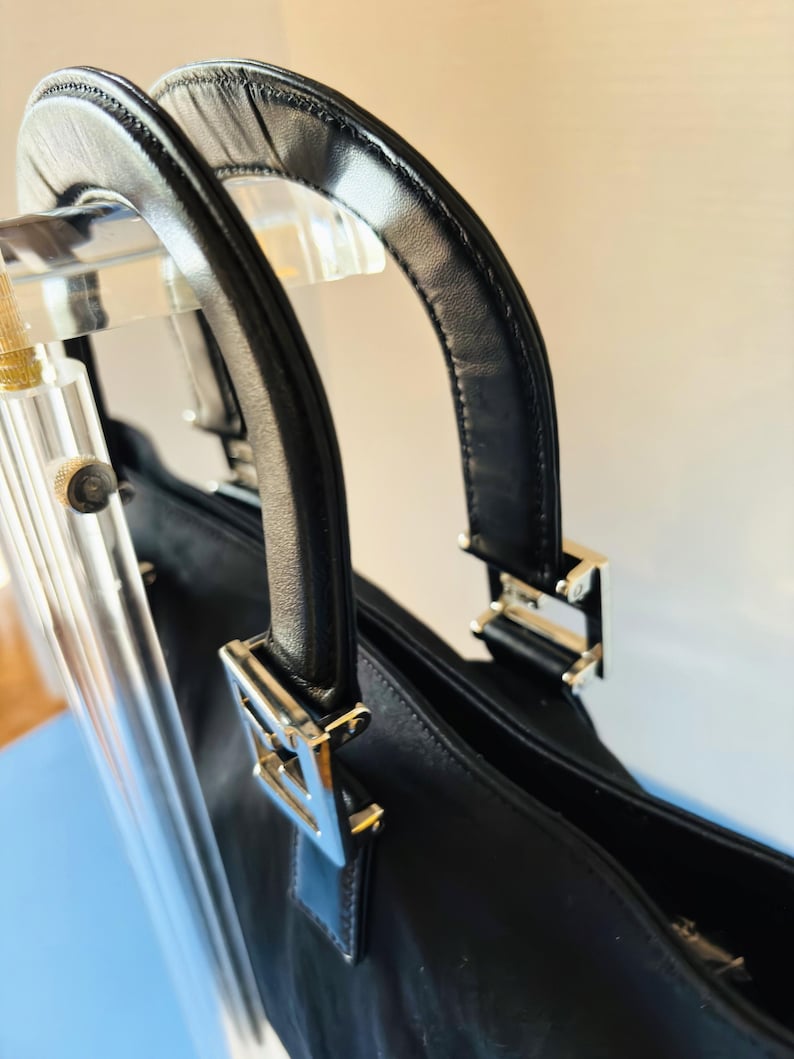 Op de afbeelding: Zwarte handtas met twee afgeronde leren handvatten en zilverkleurige metalen hardware. De handvatten van de tas zijn bevestigd aan de body met rechthoekige metalen stukken. De tas lijkt gemaakt van een glad, mogelijk lederachtig materiaal.