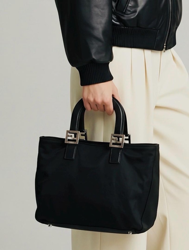 Op de afbeelding: Zwarte tote bag met zilverkleurige hardware. De tas heeft een rechthoekige vorm met twee handvatten aan de bovenkant en een stoffen body. De handvatten zijn bevestigd met zilverkleurige metalen hardware. De tas wordt door een persoon vastgehouden.