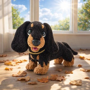 May include: A crocheted dachshund dog in black and tan. The dog has large, floppy ears and a gold chain collar. It is positioned on a wooden floor scattered with leaves, with a window and sunlight visible in the background. The dog's expression is smiling.