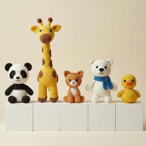 Puede incluir: Colección de cinco juguetes de animales de ganchillo hechos a mano. Incluye un panda, una jirafa, un gato, un oso polar con una bufanda azul y un pato. Los juguetes se exhiben sobre bloques blancos.