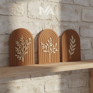Op de afbeelding: Drie terracotta-kleurige wandkunstwerken met gewelfde toppen en verticale groeven. Elk stuk heeft een ander off-white botanisch ontwerp. De kunstwerken worden tentoongesteld op een houten plank tegen een witte bakstenen muur.