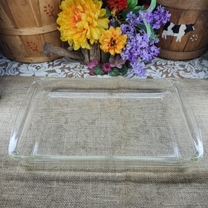 May include: A rectangular, clear glass baking dish with rounded corners. The dish is empty and sits on a burlap surface, with colorful flowers and wooden baskets in the background. The dish is approximately 13 inches long and 9 inches wide.