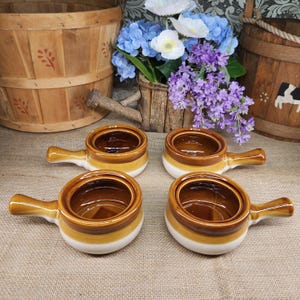 May include: Four ceramic bowls with handles, each featuring a two-tone design with a dark brown interior and rim transitioning to a lighter tan and cream base. The bowls are arranged in a square formation, showcasing a smooth, glazed finish.