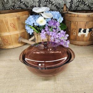 May include: A round, translucent brown glass casserole dish with a matching lid and integrated side handles. The dish features a smooth, polished surface and a circular knob on the center of the lid, reflecting light across its curved, deep bowl shape.