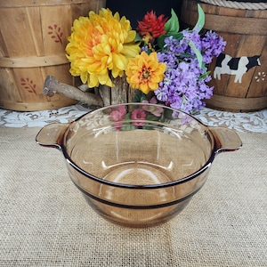 May include: A clear, amber-colored glass bowl with two handles. The bowl has a rounded shape and a dark brown rim. The background includes wooden baskets and colorful flowers, suggesting a rustic or vintage theme.