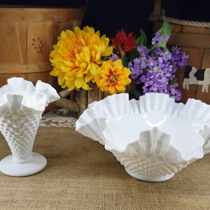 May include: Two white hobnail milk glass pieces: a ruffled-edge vase and a ruffled-edge bowl. The vase is cone-shaped with a round base. The bowl has a wide, ruffled rim. The background includes flowers and wooden baskets.