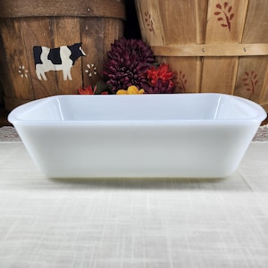 May include: A rectangular, white baking dish with slightly curved sides. The dish is made of a solid, opaque material and sits on a white surface. The background includes wooden baskets and floral accents.