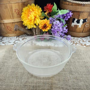 May include: A clear glass bowl with two handles. The bowl is round and has a slightly frosted appearance. The background includes wooden barrels, colorful flowers, and a burlap surface.