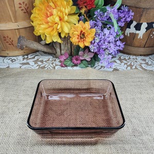 May include: A rectangular, brown glass baking dish with a black rim. The dish is empty and sits on a burlap surface. The background includes colorful flowers and a wooden basket.