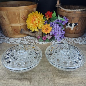 May include: Two clear, oval-shaped glass candy dishes with lids. The dishes have a textured, bubble-like pattern. The background includes wooden baskets, flowers, and a burlap surface.