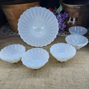 May include: A collection of white, scalloped-edge glass serving bowls. The set includes a large bowl and several smaller bowls, all with a ribbed design. The bowls are arranged on a neutral-toned surface, suggesting a vintage or antique aesthetic.