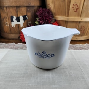May include: White mixing bowl with a blue floral design. The bowl has a silver rim and a handle. The bowl is sitting on a light-colored tablecloth. The background includes wooden baskets and red flowers.